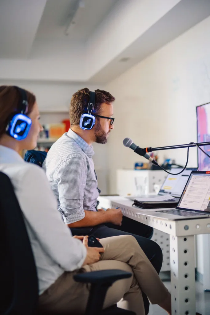 Dwie osoby siedzą przed komputerem ze słuchawkami na uszach, jedna dodatkowo przed mikrofonem.